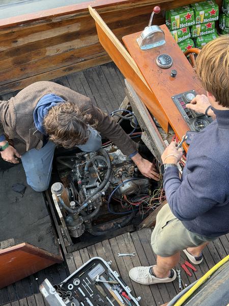 Two men are diligently working on a boat engine, surrounded by tools and wooden boat details